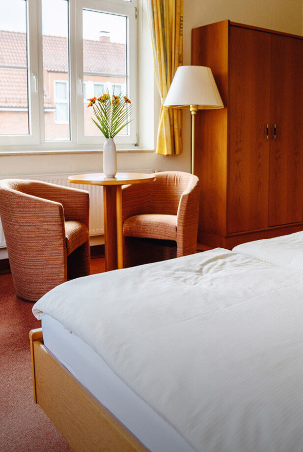 Doppelzimmer mit einem großen Bett, einem runden Holztisch, zwei Polsterstühlen, einer Stehlampe und einem Schrank. Auf dem Tisch steht eine Vase mit orangefarbenen Blumen.